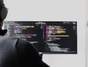Person wearing headphones sits at a desk, facing a monitor displaying code, with a desk lamp nearby.