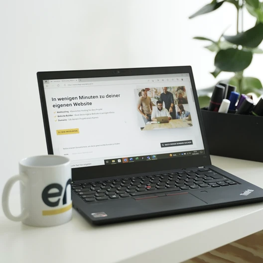 Open laptop on a white desk displaying a German website; a logo mug on the left and a black marker organizer on the right.
