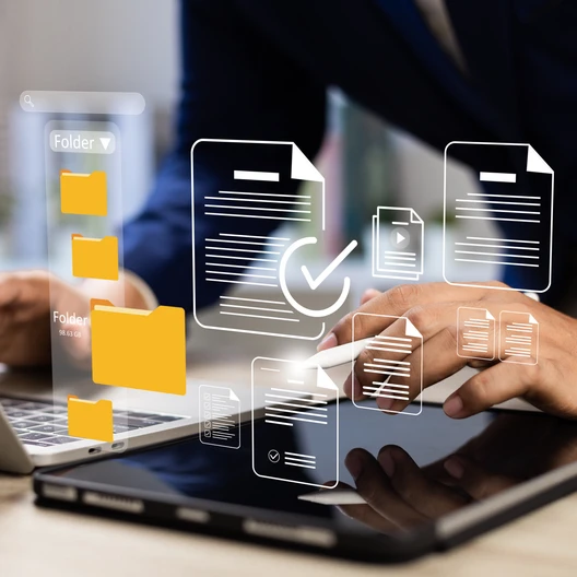 A person in a suit uses a laptop and tablet on a desk, with floating document and folder icons and a checkmark.