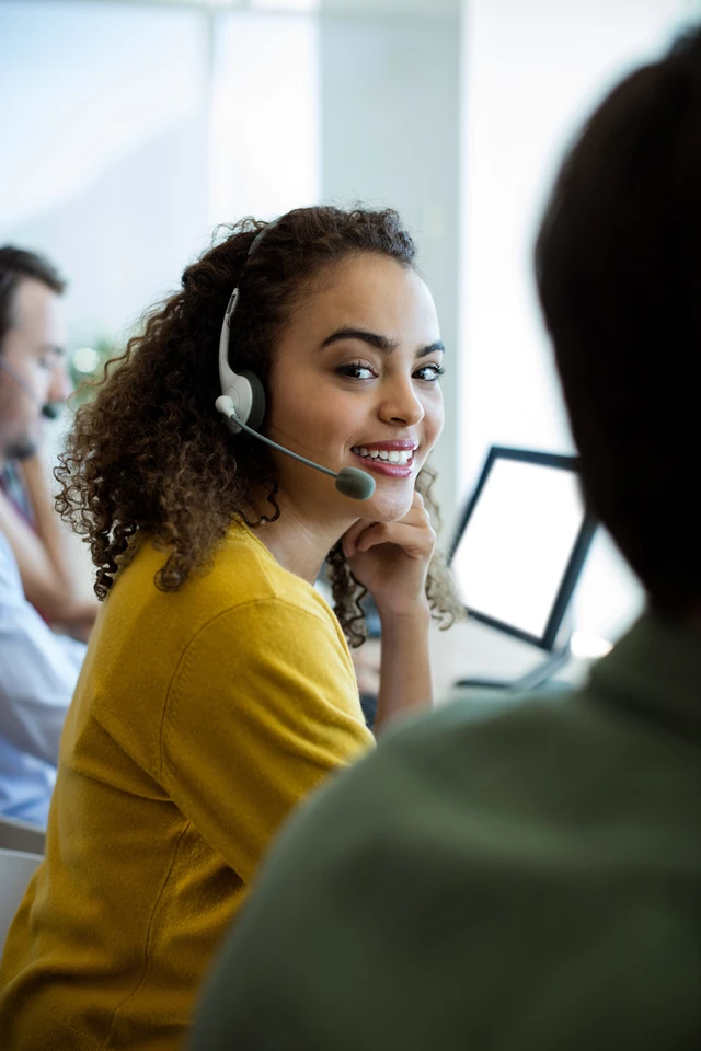 Smiling woman with curly hair wearing a headset and a yellow sweater, seated at a desk with a monitor.