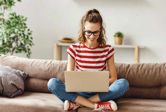 Person with striped red shirt and glasses sits cross-legged on a sofa, using a laptop.