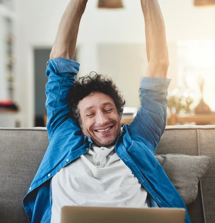 Man on a couch with a laptop, arms raised above head, smiling.