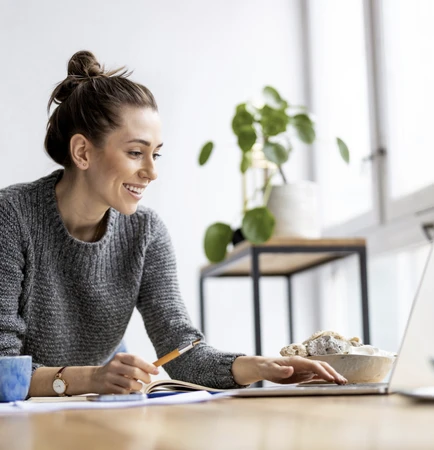 Woman with a bun in a gray sweater sits at a desk with a laptop, notebook, pencil, and a plate of cookies.