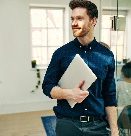 A man in a dark blue shirt holds a silver laptop under his arm in a bright office with glass walls.
