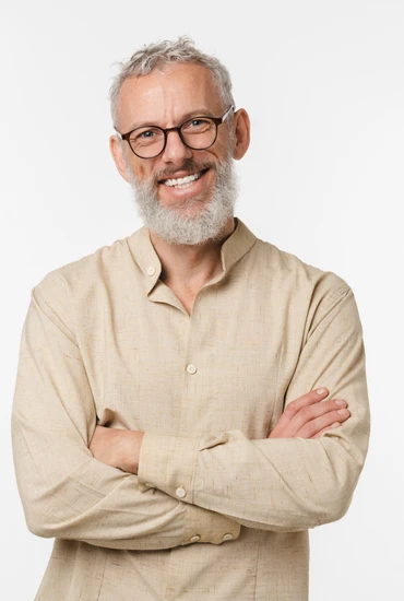 A gray-bearded man with glasses, arms crossed, wearing a beige shirt, standing against a white background.