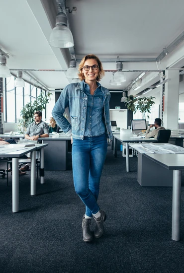 Woman in a denim jacket and jeans stands in a modern open-plan office, smiling, with desks and computers.
