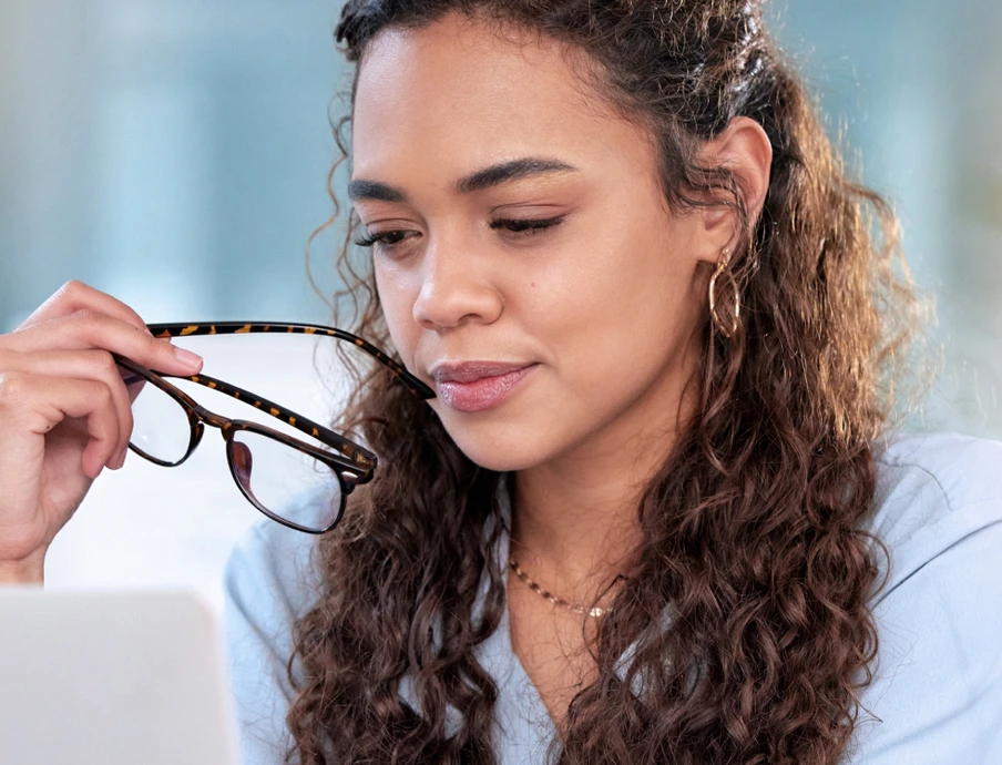 Woman with curly hair holds eyeglasses near her face, with a laptop visible in front.