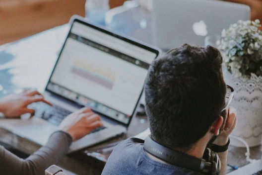 People sit at a table with open laptops; a person with a headset faces away, and a potted plant is visible.