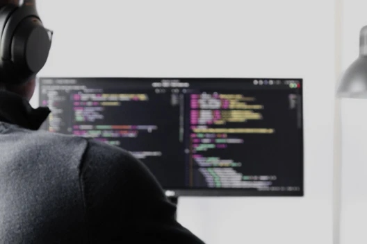 Person wearing headphones sits at a desk, facing a monitor displaying code, with a desk lamp nearby.